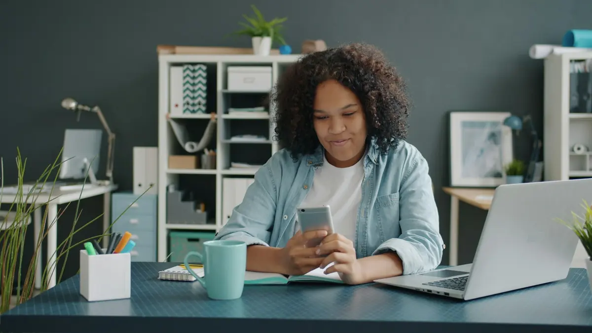 Une jeune femme consulte son téléphone à son bureau, devant un ordinateur portable